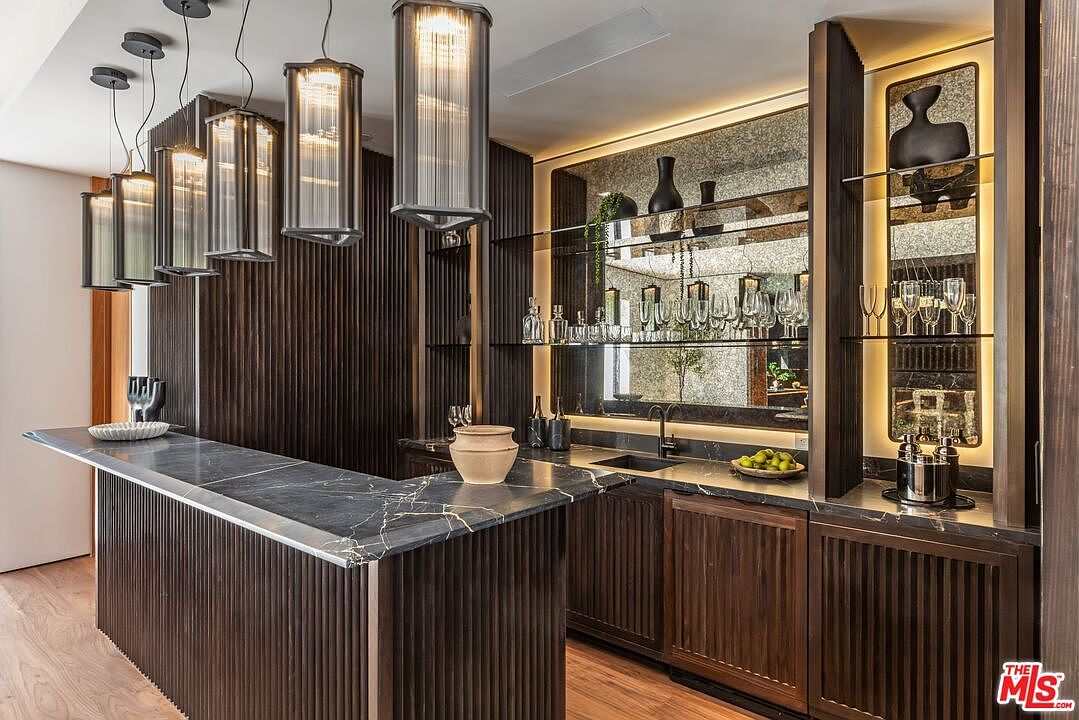 This sophisticated home bar area features a striking dark marble countertop with gold veining, complemented by rich, vertical-slatted wood cabinetry. The space is illuminated by a series of elegant, modern pendant lights and integrated under-cabinet lighting that highlights the mirrored backsplash and glass shelving. The overall aesthetic is one of refined luxury and modern design, perfect for entertaining.