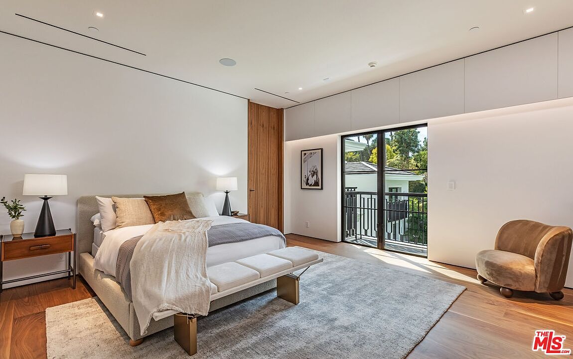 This sophisticated primary bedroom features a minimalist design with warm wood accents, a plush upholstered bed, and a neutral color palette. A large glass door leads to a private balcony, allowing natural light to flood the space, while a cozy velvet armchair provides a comfortable seating area. The room exudes a serene and luxurious atmosphere, perfect for a high-end residential listing.