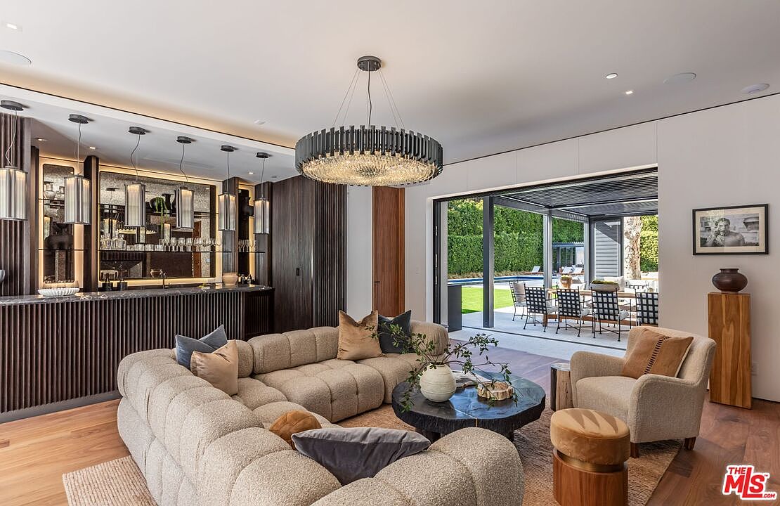 This sophisticated living room features a plush, modular sectional sofa and a unique, dark-stained live-edge coffee table, all anchored by a striking, large-scale circular chandelier. The space seamlessly transitions to an outdoor patio through expansive sliding glass doors, highlighting a wet bar with vertical wood paneling and mirrored accents. The overall aesthetic is one of modern luxury, blending warm textures with clean, contemporary lines.