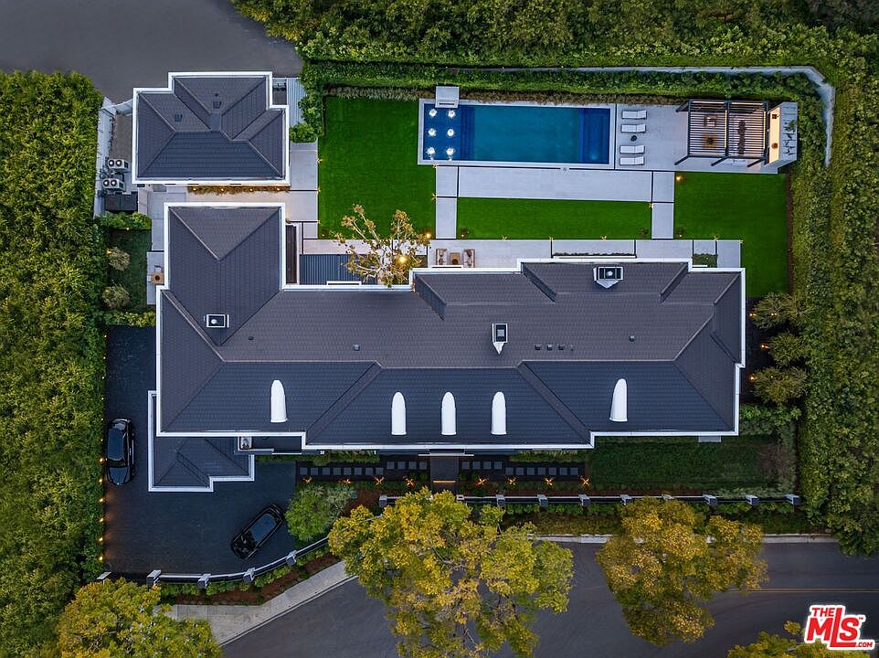 This high-angle aerial view captures a luxurious, modern estate featuring a dark-tiled, multi-gabled roof and a meticulously landscaped property. The backyard includes a rectangular swimming pool, a covered patio area, and lush green lawns, all enclosed by dense, manicured hedges for privacy. The property is situated on a corner lot with a paved driveway and mature trees, presenting an elegant and secluded residential atmosphere.
