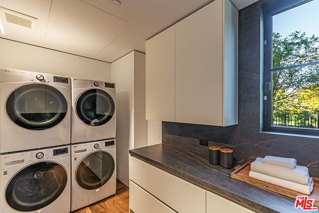 This modern, high-end laundry room features a stacked configuration of four white LG front-load washers and dryers, maximizing space efficiency. The room is accented by sleek, dark stone-look countertops and matching backsplash, complemented by minimalist white cabinetry and a window that brings in natural light. The overall aesthetic is clean, organized, and sophisticated, reflecting a contemporary design sensibility.