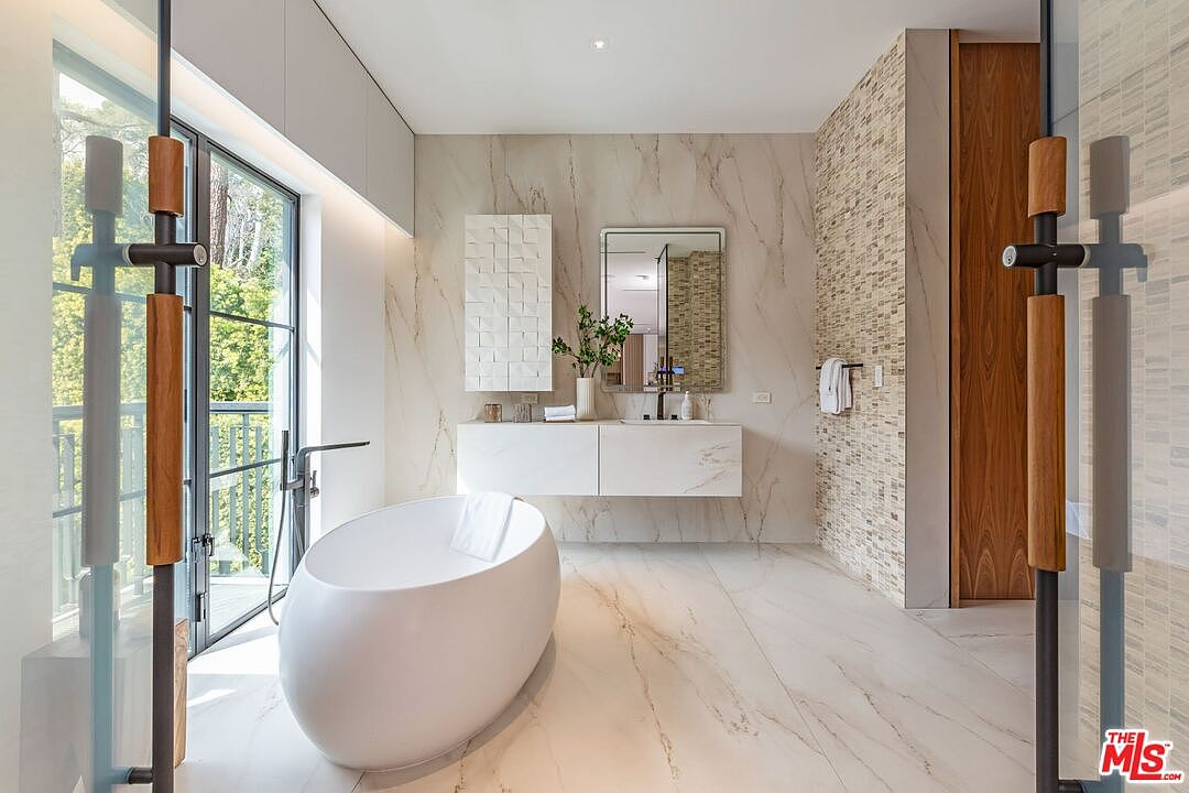 This luxurious primary bathroom features a sleek, freestanding white soaking tub positioned next to floor-to-ceiling windows that offer a serene view of the surrounding greenery. The space is anchored by a floating white vanity with marble-veined walls and a textured stone accent wall, creating a sophisticated, spa-like atmosphere. The perspective is centered, showcasing the clean lines and high-end finishes that define this modern, elegant retreat.