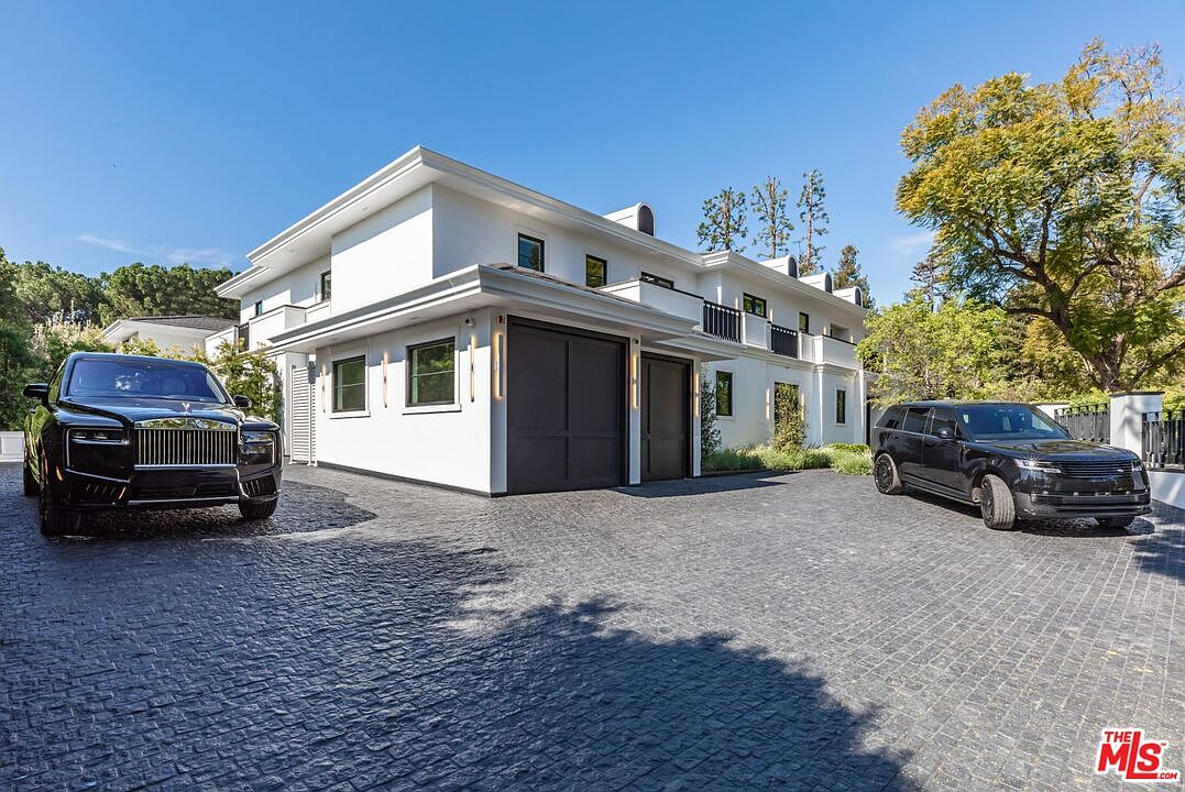 This image showcases the front exterior of a modern, white-stucco luxury residence featuring a prominent two-car garage with dark, paneled doors. A cobblestone driveway leads up to the house, flanked by two high-end black SUVs, creating an impression of exclusivity and wealth. The architectural style is clean and contemporary, with balanced proportions, black-framed windows, and a second-story balcony, all set against a clear blue sky.
