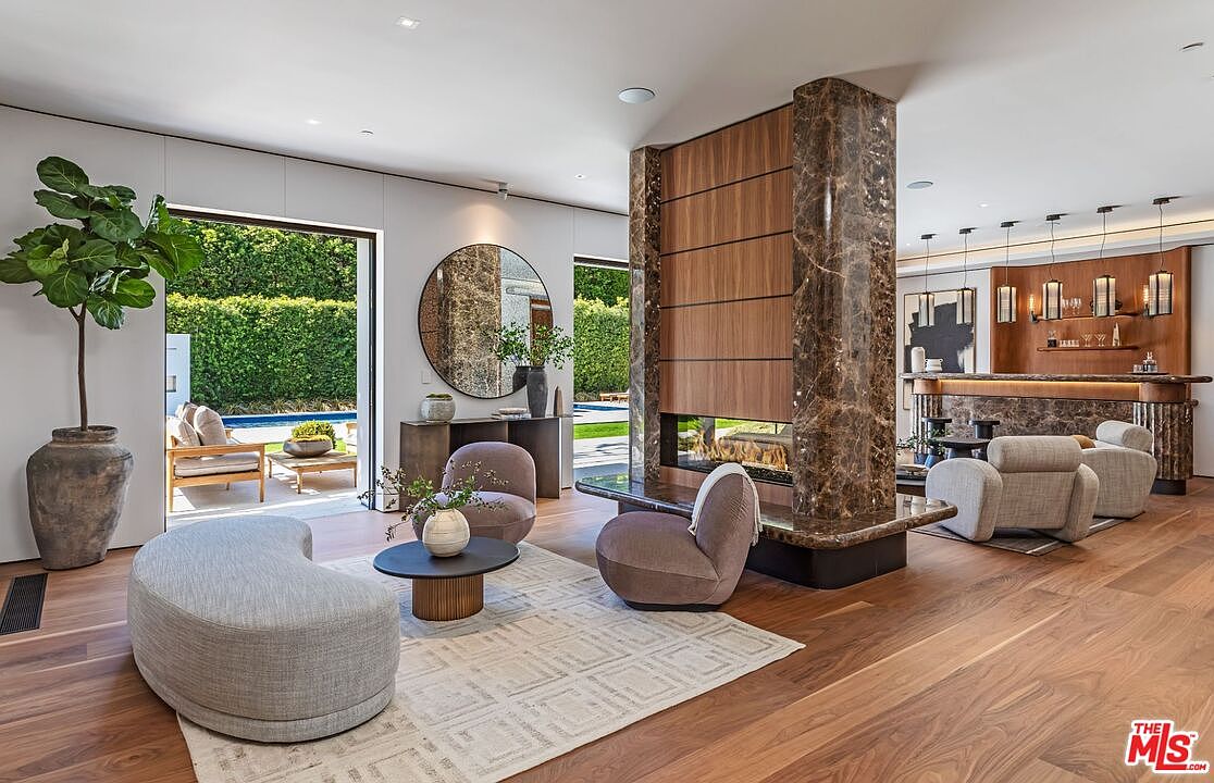 This sophisticated open-concept living area features a striking floor-to-ceiling fireplace clad in rich brown marble and wood paneling, serving as a central architectural anchor. The space is furnished with plush, curved seating in neutral tones, a textured area rug, and a minimalist coffee table, all set against warm hardwood flooring. Large sliding glass doors seamlessly connect the interior to a sun-drenched patio and pool area, creating an airy, luxurious indoor-outdoor flow.