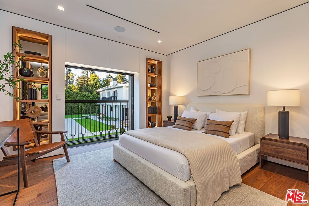 This sophisticated primary bedroom features a neutral color palette, a plush upholstered bed, and warm wood flooring, creating a serene and luxurious atmosphere. Large sliding glass doors open onto a private balcony, allowing for abundant natural light and a seamless connection to the outdoors, while built-in illuminated shelving adds a custom architectural touch. The room is elegantly appointed with modern bedside lamps and minimalist decor, emphasizing a clean, contemporary aesthetic.