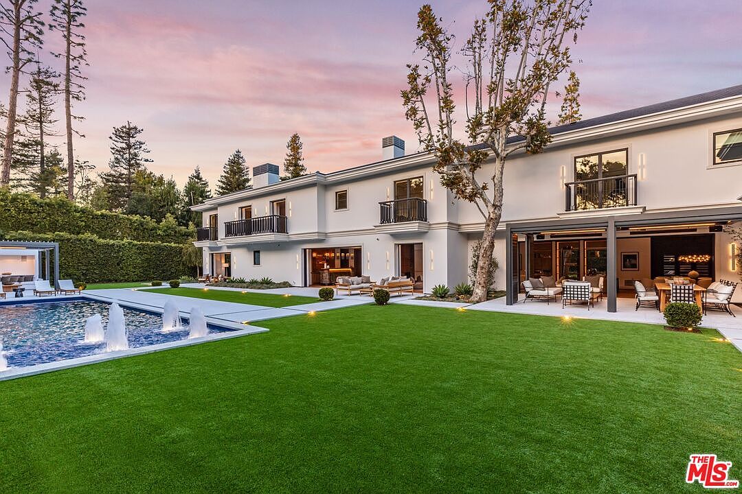 This expansive rear view of a luxury estate showcases a pristine, manicured lawn leading to a modern, multi-story residence with a large swimming pool featuring integrated fountains. The home features a contemporary architectural style with clean lines, multiple balconies, and a covered outdoor patio area perfect for entertaining. The scene is captured during a vibrant sunset, casting a warm, inviting glow over the entire property.