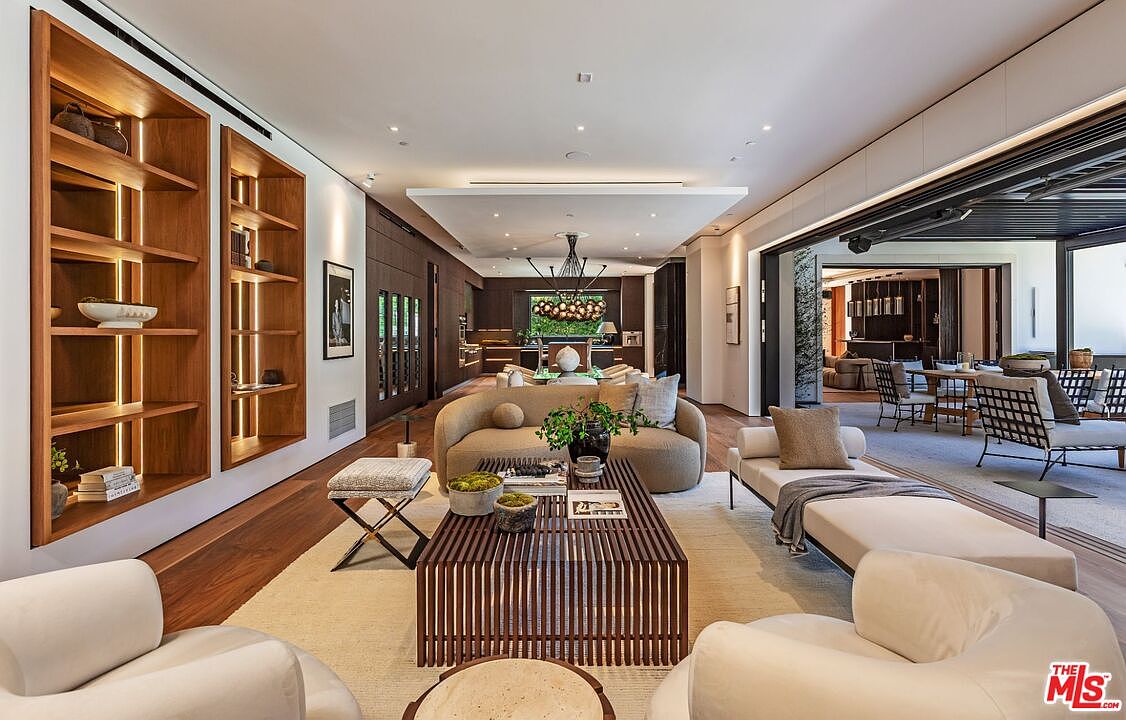 This expansive, open-concept living room exudes modern luxury with its warm wood accents, recessed lighting, and a seamless transition to an outdoor patio through large sliding glass doors. The space is anchored by a slatted wooden coffee table and a curved sofa, creating a sophisticated yet inviting atmosphere. The perspective captures the depth of the home, showcasing a cohesive flow from the living area toward the dining space and kitchen in the background.
