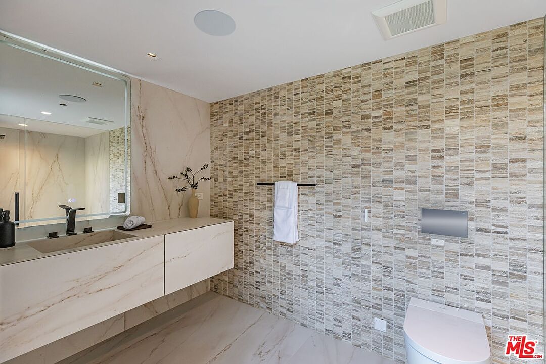 This modern primary bathroom features a sleek, wall-mounted vanity with a marble countertop and integrated sink, complemented by a large, frameless mirror. The space is defined by a striking textured stone accent wall that adds depth and warmth, contrasting with the smooth, light-toned marble flooring. A wall-mounted toilet and minimalist fixtures complete the sophisticated, high-end aesthetic of this contemporary bathroom.