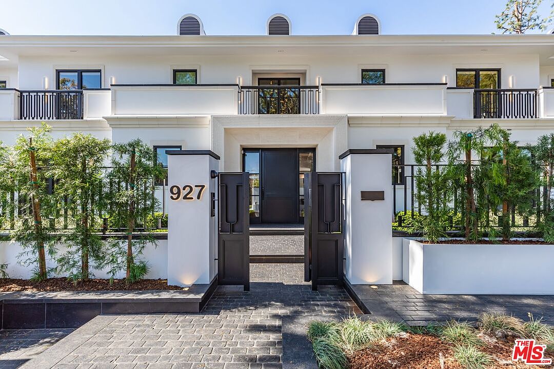 This striking modern entryway features a symmetrical white facade with clean lines and a prominent black metal gate leading to the front door. The property is accented by manicured landscaping, including tall, slender trees and white planters, creating a sophisticated and welcoming curb appeal. The perspective is a direct, eye-level shot that emphasizes the grand, contemporary architectural design of the home.