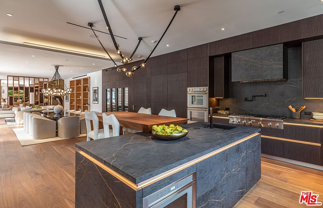 This sophisticated, modern kitchen features dark, textured cabinetry paired with dramatic, veined stone countertops and a matching waterfall island. The open-concept design seamlessly flows into a dining area, highlighted by a striking, sculptural chandelier and warm wood flooring. The space exudes luxury with its high-end built-in appliances, integrated lighting, and a clean, minimalist aesthetic.
