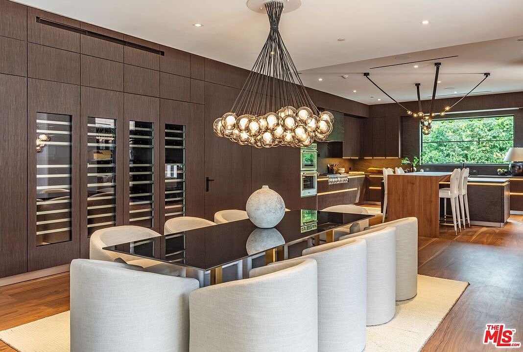 This sophisticated dining room features a sleek, dark-toned aesthetic with a large, reflective dining table surrounded by plush, light-colored upholstered chairs. A striking, multi-bulb chandelier serves as the focal point above the table, while integrated wine storage cabinets with glass doors line the wall. The open-concept design seamlessly transitions into a modern kitchen, creating an elegant and cohesive space perfect for high-end entertaining.