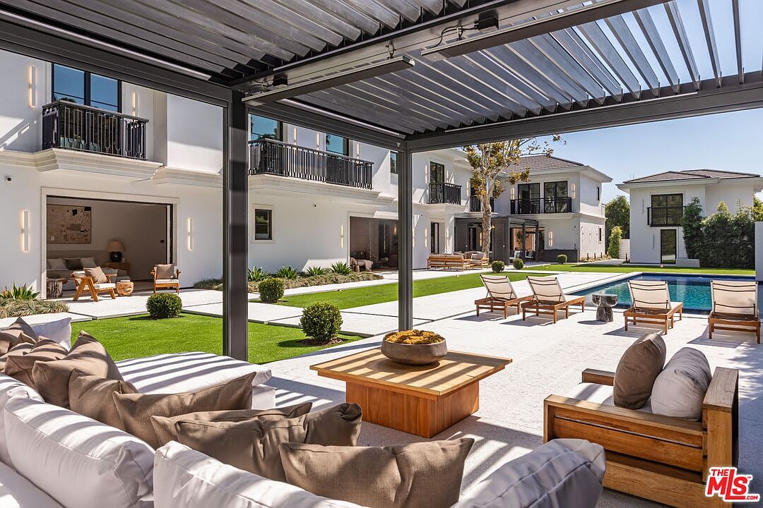 This luxurious outdoor living space features a modern pergola covering a comfortable lounge area with plush seating and a wooden coffee table. The patio seamlessly transitions into a manicured lawn and a sparkling swimming pool, framed by the elegant white architecture of the multi-story estate. The perspective from under the pergola creates an inviting, cinematic frame that highlights the sophisticated blend of indoor-outdoor living.
