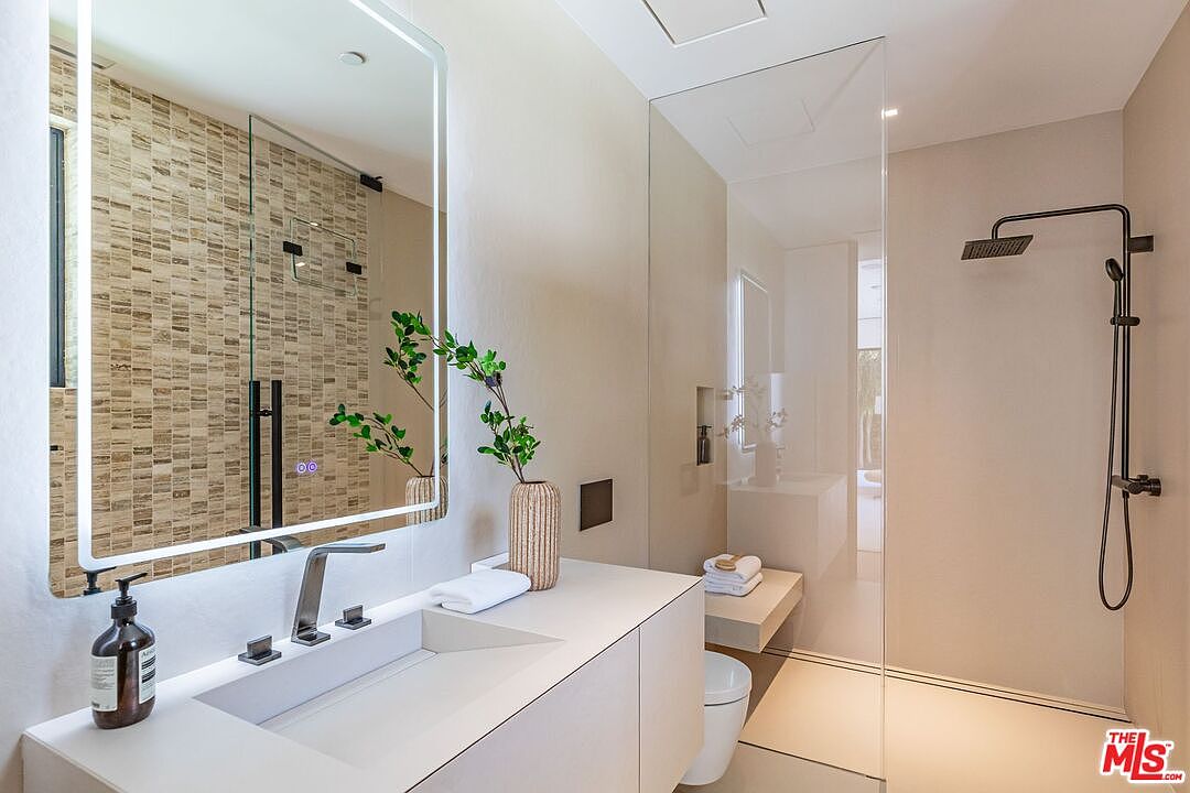 This modern primary bathroom features a sleek, minimalist vanity with a white countertop and a large, illuminated rectangular mirror. To the right, a spacious walk-in shower is enclosed by a frameless glass panel, showcasing a textured stone accent wall and a contemporary rainfall showerhead. The overall aesthetic is clean, sophisticated, and spa-like, emphasizing high-end finishes and neutral tones.