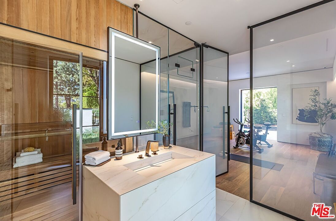 This luxurious primary bathroom features a modern floating vanity with a marble countertop and a sleek, illuminated mirror. To the left, a glass-enclosed sauna with warm wood paneling adds a spa-like element, while a glass partition separates the space from an adjacent home gym. The design is sophisticated and contemporary, utilizing high-end materials and an open-concept layout that seamlessly integrates wellness features.