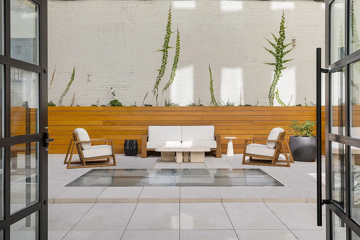 This is an inviting outdoor patio space. It features modern furniture, including a white sofa and armchairs, arranged around a low table. The space is decorated with climbing greenery on a white brick wall and potted plants, creating a serene and stylish atmosphere.