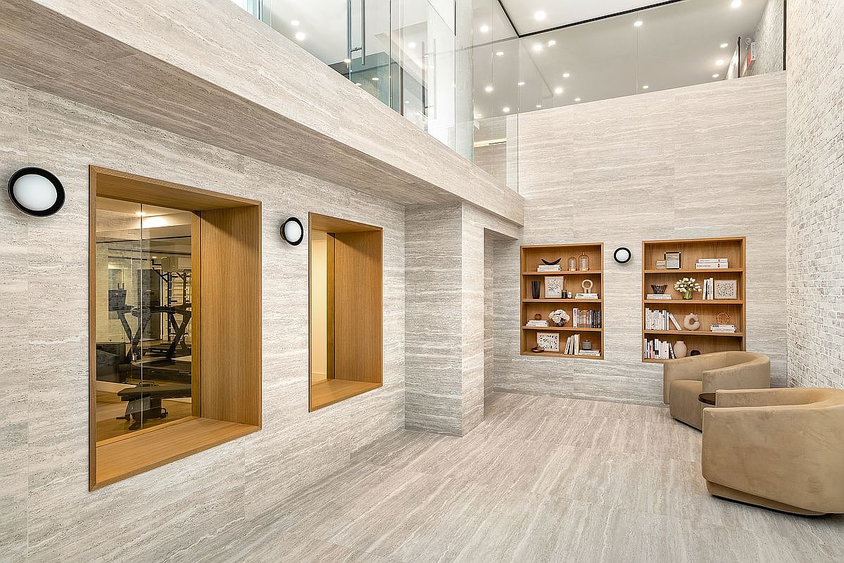 This interior shot showcases a modern hallway with travertine walls and flooring. The hallway features built-in bookshelves, recessed lighting, and framed openings, one of which reveals a home gym. The space is well-lit and exudes a sense of luxury and sophistication.