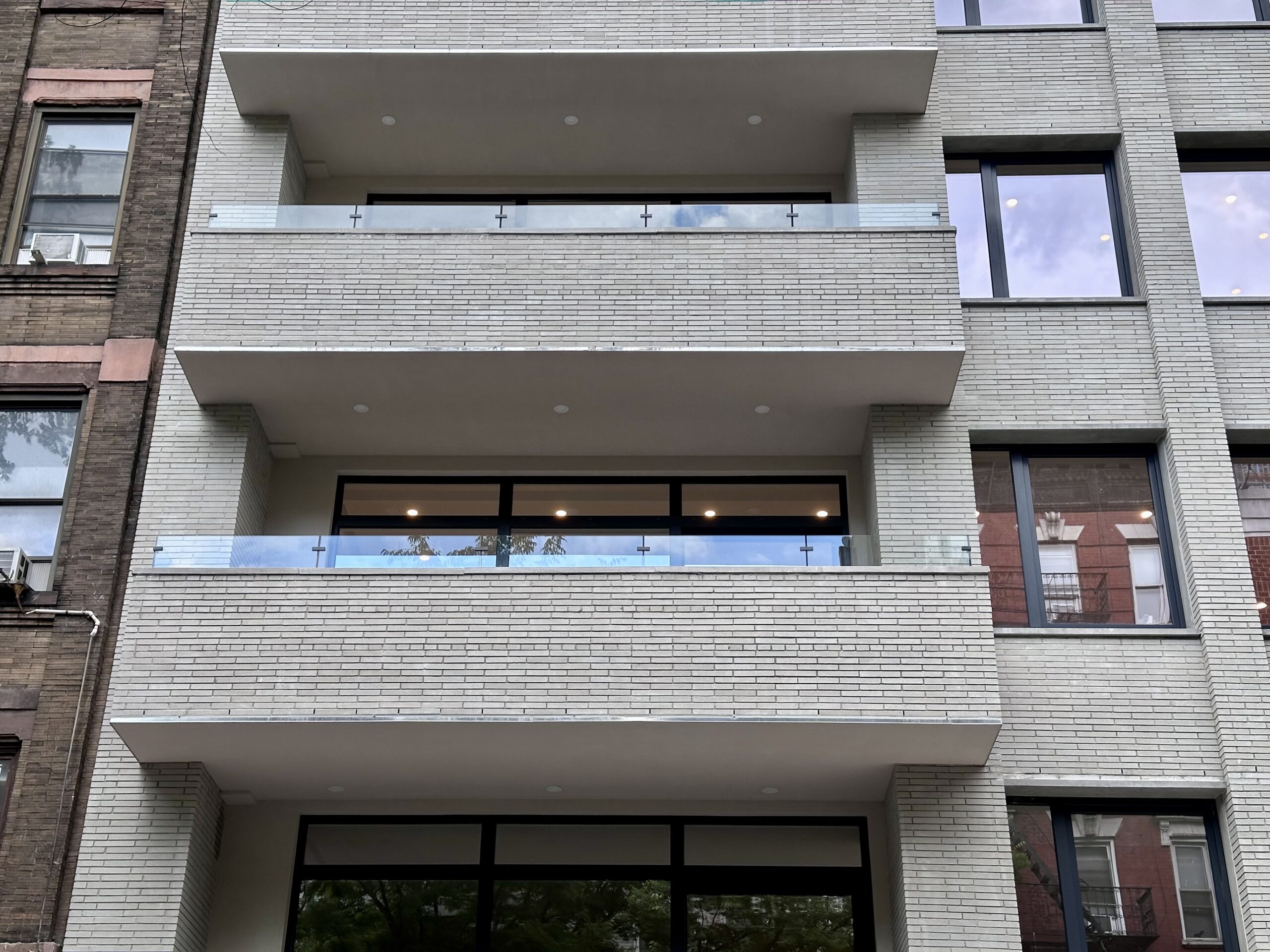 This is a front view of an apartment building showcasing modern architectural details. The facade features light-colored brickwork with protruding balconies enclosed by glass railings. The windows reflect the sky and surrounding buildings, indicating a contemporary urban setting.