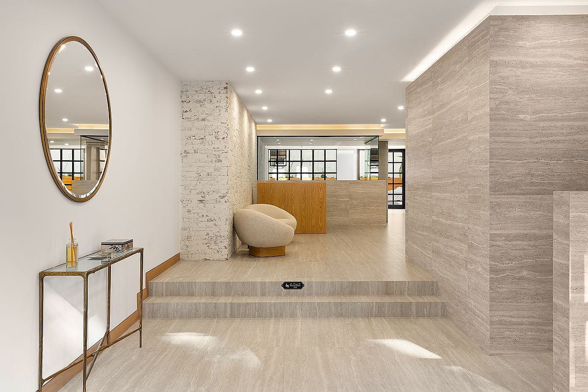 This interior shot showcases a modern hallway with a minimalist design. The space features a light-colored stone floor, a textured wall, and a decorative mirror above a console table. The hallway leads to a reception area with a wooden desk and a comfortable chair, creating a welcoming and sophisticated atmosphere.