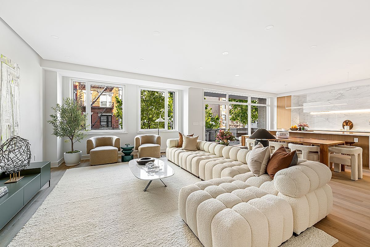 This is a bright and spacious living room featuring a large, modular sofa with a textured, off-white fabric. The room is well-lit with natural light from large windows, offering views of the outdoors. The decor is modern and minimalist, with a neutral color palette and a focus on clean lines and comfortable furnishings, creating an inviting and luxurious atmosphere.