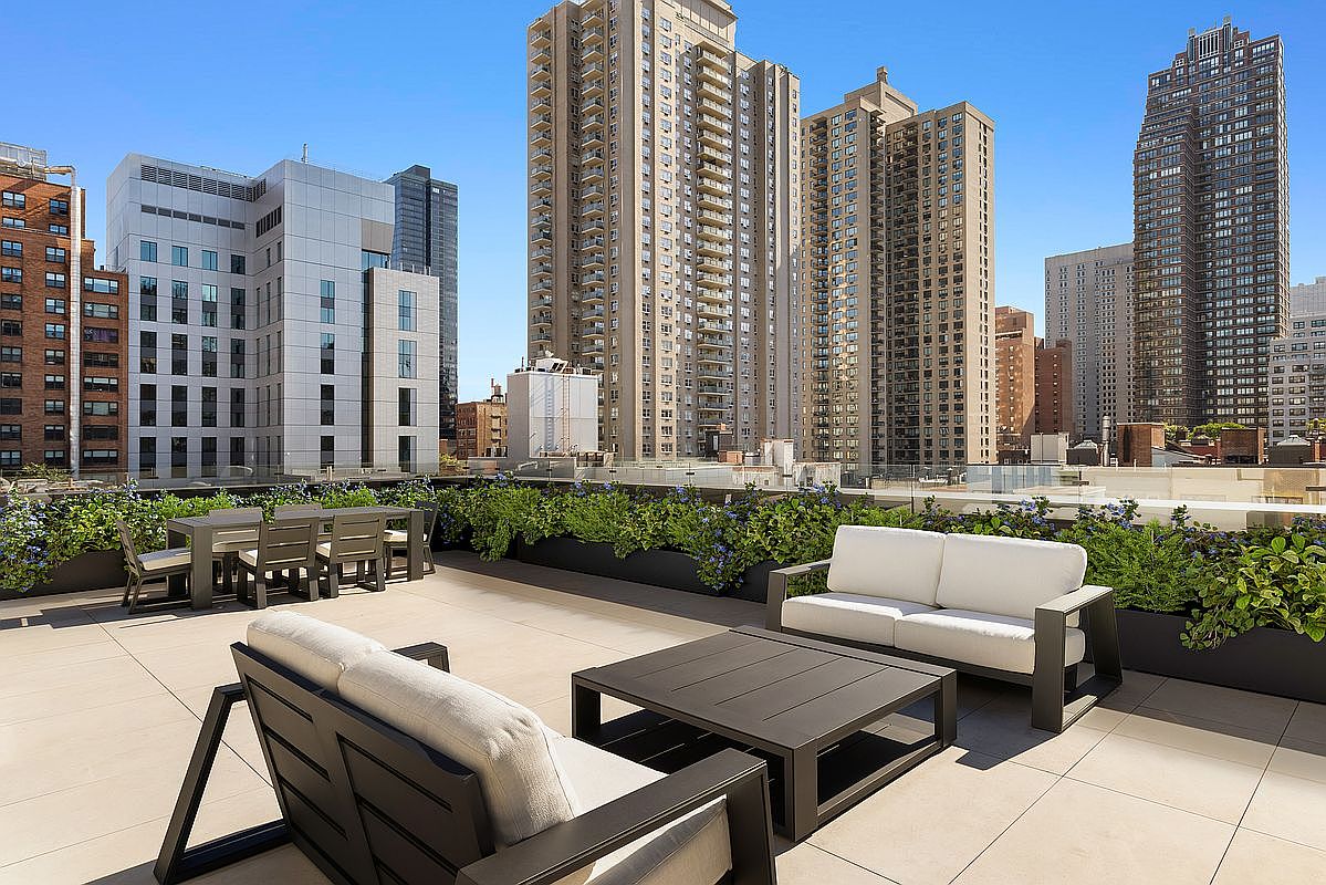 This image showcases a luxurious rooftop patio or deck area, furnished with modern outdoor seating including a sofa, chairs, and a coffee table. Lush greenery lines the perimeter, providing a sense of privacy and tranquility amidst the urban backdrop of tall buildings under a clear blue sky. The space is designed for relaxation and entertainment, offering a sophisticated outdoor living experience.
