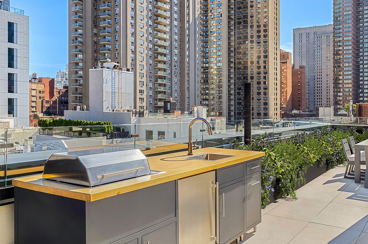 This image showcases a luxurious outdoor kitchen on a patio or balcony, featuring a built-in grill, sink, and storage cabinets with a sleek, modern design. The countertop is a warm wood tone, contrasting with the gray cabinetry. The backdrop includes a cityscape with tall buildings, suggesting an urban setting, and greenery adds a touch of nature to the space.