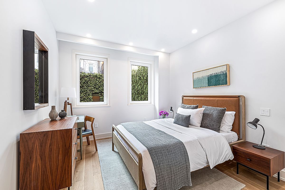 This is a well-lit bedroom featuring a bed with a light wood frame and a leather headboard, adorned with white linens and gray pillows. A wooden dresser with a mirror sits against the wall, complemented by a small desk and chair. Two windows offer views of greenery, enhancing the room's bright and airy atmosphere.