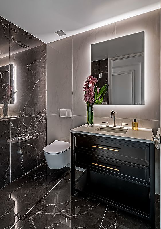 This is a modern primary bathroom featuring dark marble walls and flooring with white veining, creating a luxurious and sophisticated ambiance. A sleek black vanity with gold hardware is topped with a white countertop and a vessel sink, complemented by a large, illuminated mirror. The toilet is a modern, wall-mounted design, and the overall impression is one of high-end elegance and contemporary style.