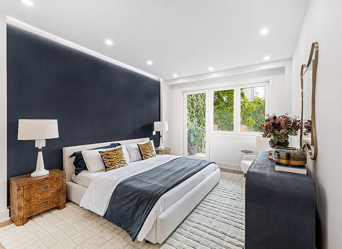 This is a well-lit primary bedroom featuring a king-size bed with a dark blue throw blanket and decorative pillows. The room has a dark blue accent wall behind the bed, recessed lighting, and a light-colored rug. A wooden nightstand with a lamp sits beside the bed, and a dark blue dresser with a mirror and floral arrangement is positioned on the opposite side of the room, creating a balanced and inviting space.