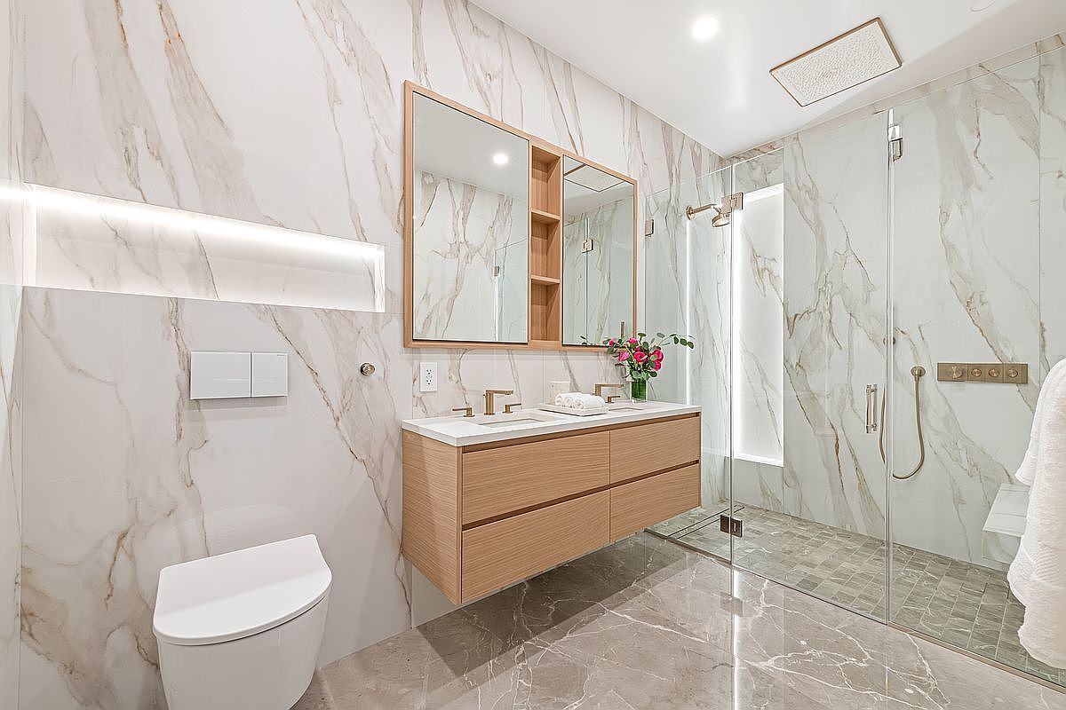 This is a modern primary bathroom featuring marble walls and flooring, a floating wood vanity with double sinks, and a glass-enclosed shower. The design is sleek and luxurious, with a focus on clean lines and high-end finishes. The perspective is from the doorway, showcasing the entire space.