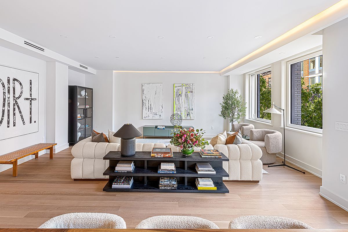 This is a bright and airy living room featuring hardwood floors, white walls, and recessed lighting. A large, abstract art piece hangs on the wall, complemented by modern furniture including a cream-colored sofa, a dark wood console table, and two armchairs near the window. The room exudes a sophisticated and comfortable atmosphere.