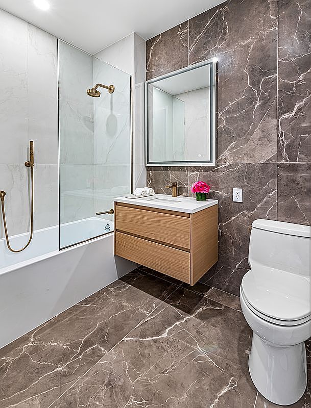This is a modern primary bathroom featuring a floating vanity with a light wood finish, a white countertop, and a square mirror with integrated lighting. The walls are tiled with a combination of white marble-look tiles in the shower area and dark brown marble-look tiles on the adjacent wall. The flooring is also a dark brown marble-look tile, creating a cohesive and luxurious aesthetic. A white toilet is visible on the right side of the image.