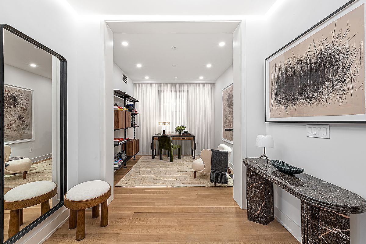 This interior shot showcases a hallway leading into a bright office space. The hallway features hardwood floors, a large framed artwork, a marble console table with decorative items, and a full-length mirror reflecting a seating area. The office beyond is well-lit with natural light filtering through sheer curtains, and contains a desk, chair, and shelving unit, creating an inviting and functional workspace.