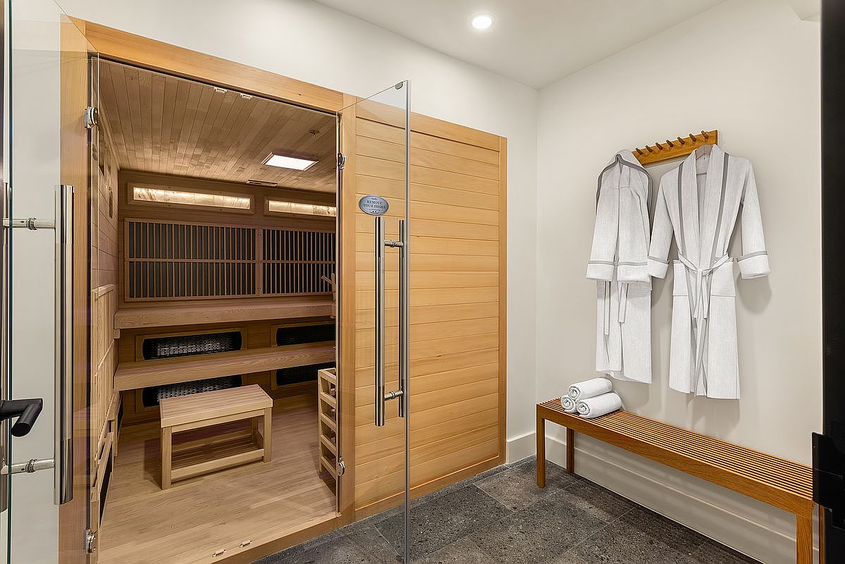 This image showcases a modern sauna and adjacent bathroom area. The sauna features wooden benches and a glass door, while the bathroom area includes a wooden bench with neatly stacked towels and two robes hanging on a rack. The flooring is a dark tile, creating a spa-like atmosphere.