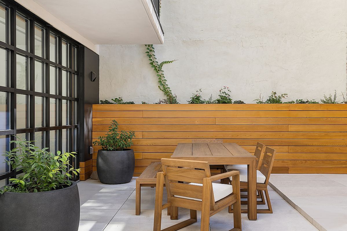 This image showcases a stylish outdoor patio area, featuring a wooden dining set with cushioned chairs and a bench, set against a backdrop of a wooden planter box with greenery. The patio is paved with large tiles, and a black-framed window wall adds a modern touch. The overall impression is one of a serene and well-designed outdoor living space.