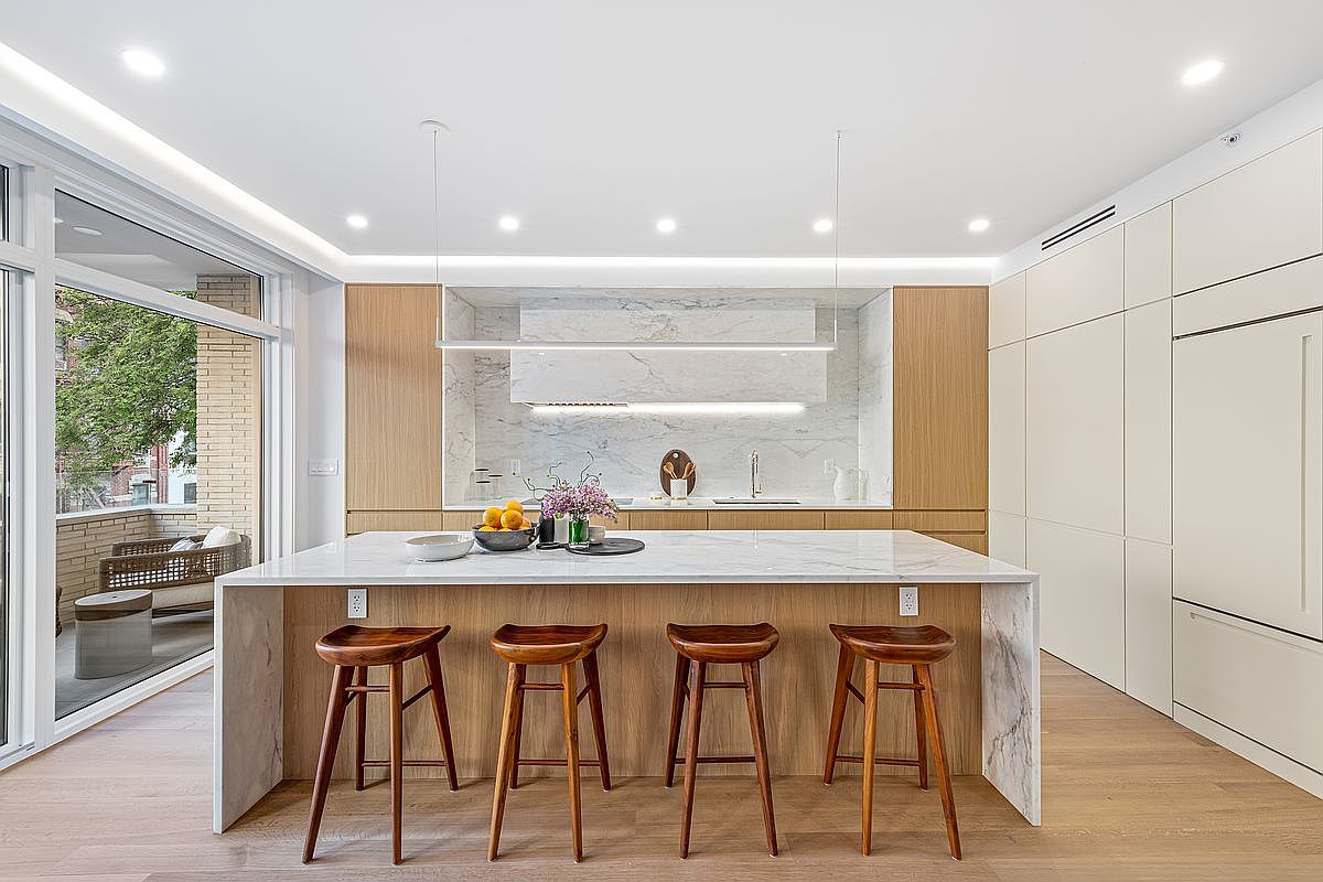 This is a bright and modern kitchen featuring a large island with a marble countertop and wooden stools. The kitchen has light wood cabinetry and a marble backsplash, complemented by sleek, linear lighting fixtures. A sliding glass door opens to an outdoor patio, enhancing the sense of space and light.