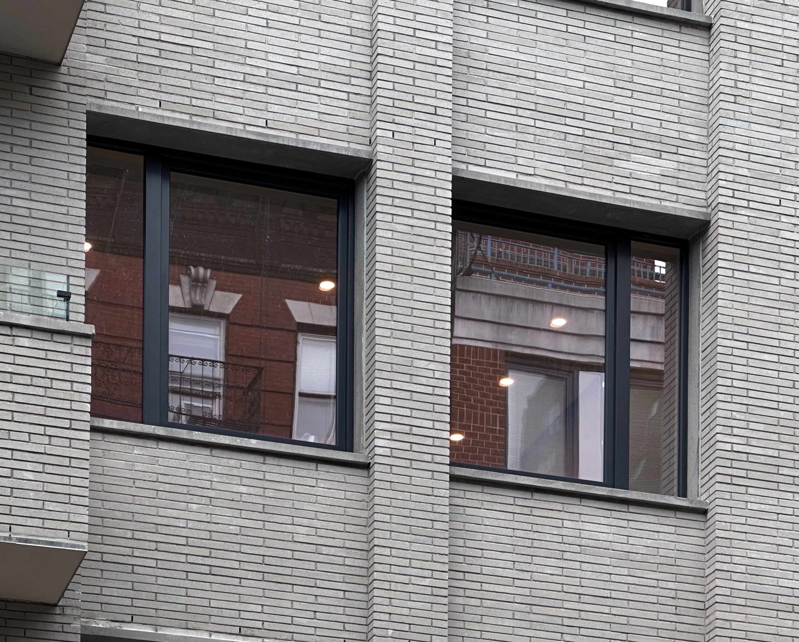 This image shows the exterior front view of a building. The facade is constructed from light gray brick, featuring clean, horizontal lines. Two large windows with dark frames reflect surrounding buildings, adding depth and interest to the architectural design.