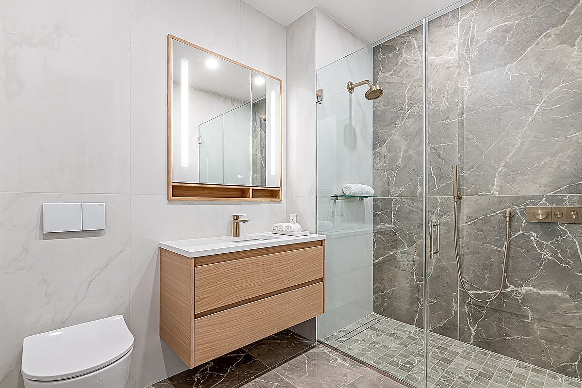 This is a modern primary bathroom featuring a floating wood vanity with a white countertop and a large, illuminated mirror above. The shower is enclosed with glass and features gray marble-like tile, along with gold fixtures. The floor is a dark marble, and the overall impression is clean and luxurious.