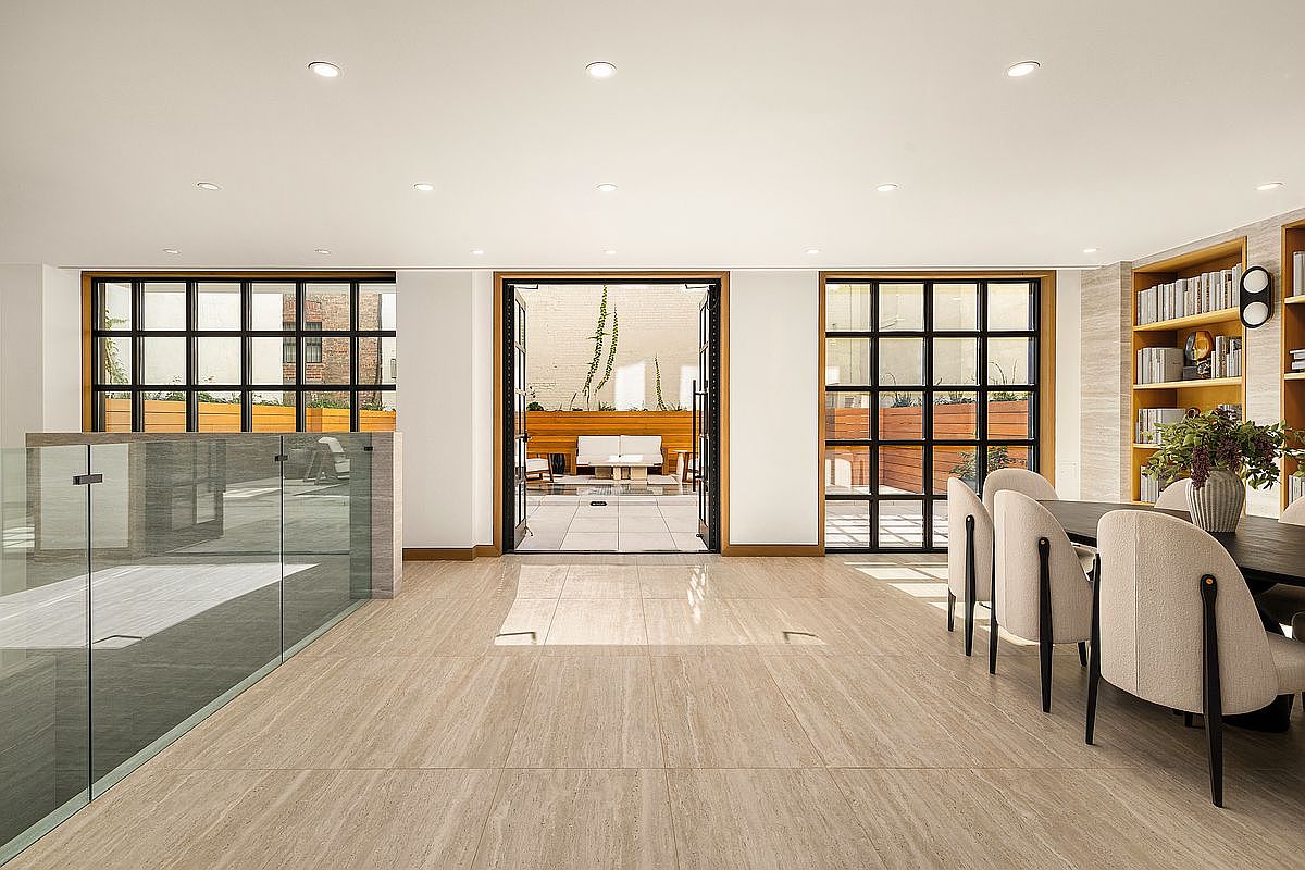 This is a bright and airy dining room featuring large windows with black frames that offer views of an outdoor patio. The room is decorated in a modern style with a neutral color palette, including light-colored flooring and dining chairs. A built-in bookshelf adds a touch of sophistication, and the open doorway leads to an outdoor space, creating a seamless indoor-outdoor flow.