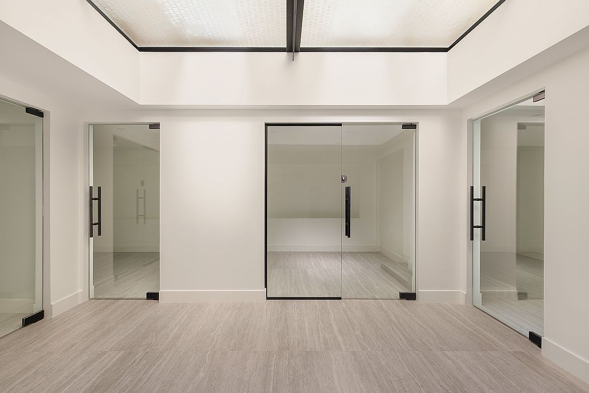 This interior shot showcases a modern hallway with a clean and minimalist design. The hallway features light gray flooring and white walls, complemented by three glass doors with black frames and handles. The ceiling has a unique grid-like pattern, adding a touch of architectural interest to the space.