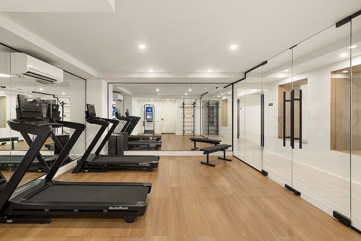 This is a well-equipped home gym featuring multiple treadmills, weight benches, and wall-mounted exercise equipment. The room has a clean and modern aesthetic with light wood flooring, mirrored walls to enhance the sense of space, and glass partitions that create a bright and airy atmosphere. The space is designed for a comprehensive workout experience.