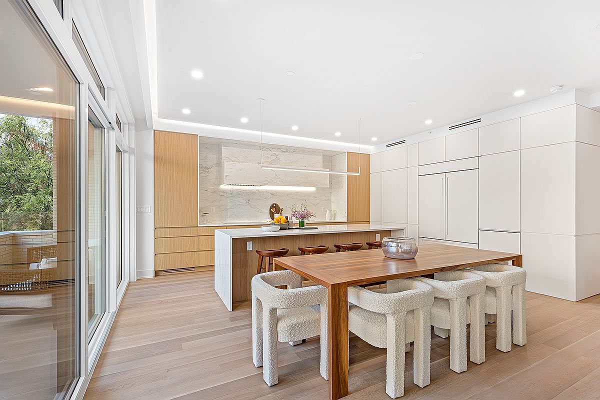 This is a bright and modern kitchen featuring light wood flooring, a large island with seating, and a dining table with unique, plush chairs. The kitchen boasts a combination of light wood and white cabinetry, complemented by a marble backsplash and sleek, linear lighting fixtures. Large windows provide ample natural light, enhancing the clean and contemporary aesthetic.