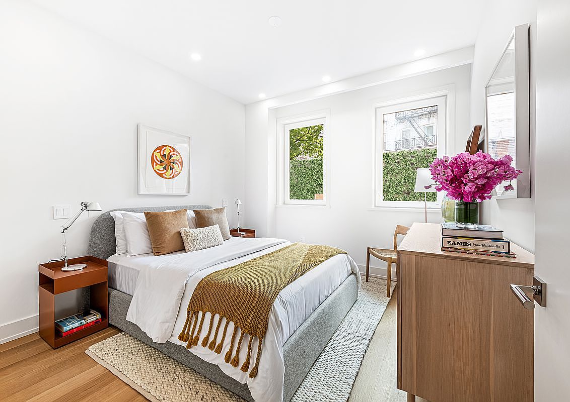 This is a bright and airy bedroom featuring a bed with a gray upholstered frame, white linens, and a mustard-colored throw. The room is decorated with a modern art piece, a wooden nightstand, and a dresser with a vase of pink flowers. Natural light streams in through two windows, creating a welcoming and comfortable atmosphere.