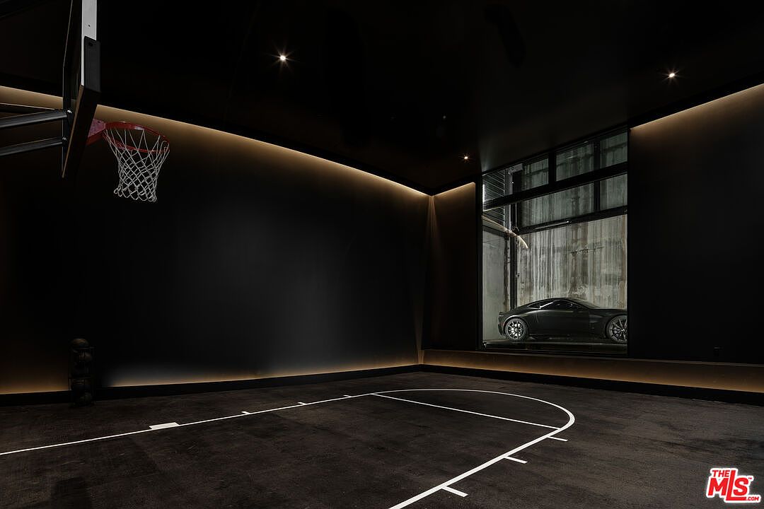 This sleek, modern indoor basketball court features a dramatic all-black aesthetic with recessed perimeter lighting that creates a sophisticated, moody atmosphere. A large glass window provides a unique, cinematic view into an adjacent garage, showcasing a luxury sports car as a design element. The space is minimalist and high-end, emphasizing a seamless blend of athletic utility and luxury automotive display.