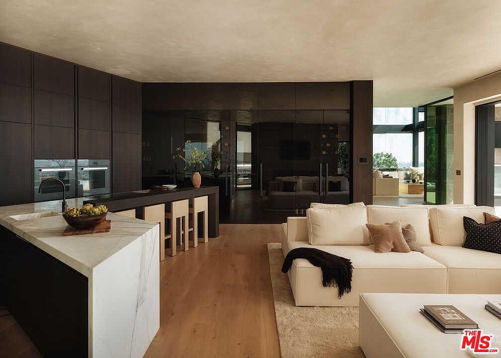 This sophisticated open-concept space seamlessly blends a modern kitchen with a cozy living area, featuring dark wood cabinetry, a marble-topped island, and light oak flooring. The kitchen is equipped with high-end built-in appliances and a breakfast bar, while the adjacent living room is anchored by a plush, cream-colored sectional sofa. The perspective captures the elegant flow of the home, highlighting the contrast between the dark, reflective surfaces and the warm, inviting textures of the living space.