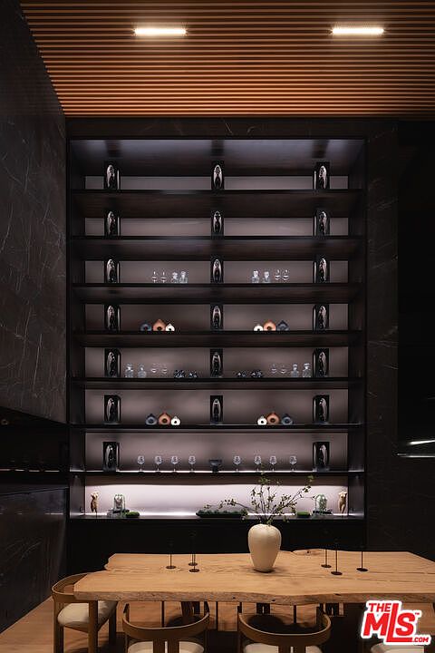 This sophisticated dining area features a live-edge wooden table as the centerpiece, complemented by modern, minimalist chairs. A striking, floor-to-ceiling dark wood shelving unit with integrated LED lighting serves as a dramatic focal point, displaying curated decorative objects and glassware. The room is defined by its moody, high-end aesthetic, enhanced by a slatted wood ceiling and dark marble-like wall accents.