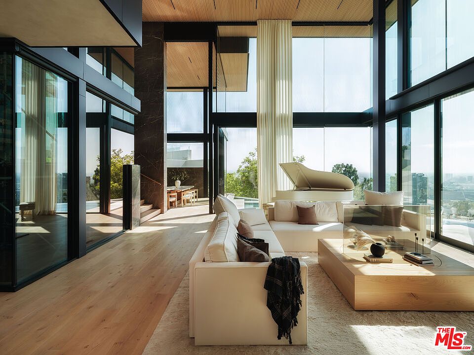 This expansive, ultra-modern living room features floor-to-ceiling glass walls that flood the space with natural light and offer panoramic views. The room is anchored by a plush, cream-colored sectional sofa, a minimalist light wood coffee table, and a grand piano, all set against a backdrop of warm wood-slatted ceilings and dark marble accents. The open-concept design seamlessly connects the living area to a dining space in the background, creating an atmosphere of sophisticated, high-end luxury.