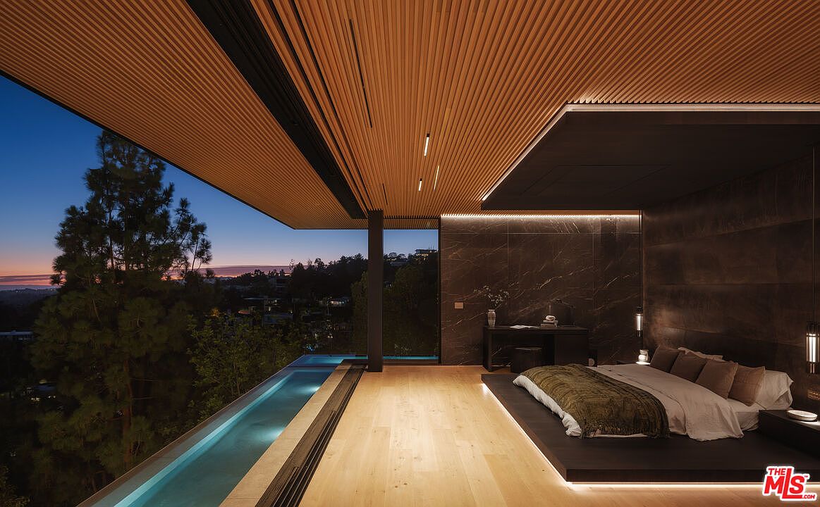 This luxurious primary bedroom features a seamless indoor-outdoor design with a private infinity-edge pool directly adjacent to the sleeping area. The room is defined by a sophisticated, minimalist aesthetic, showcasing a raised platform bed, dark marble accent walls, and a warm, slatted wood ceiling. The expansive glass wall offers a breathtaking, cinematic view of the twilight sky and surrounding landscape, creating an atmosphere of serene, high-end tranquility.