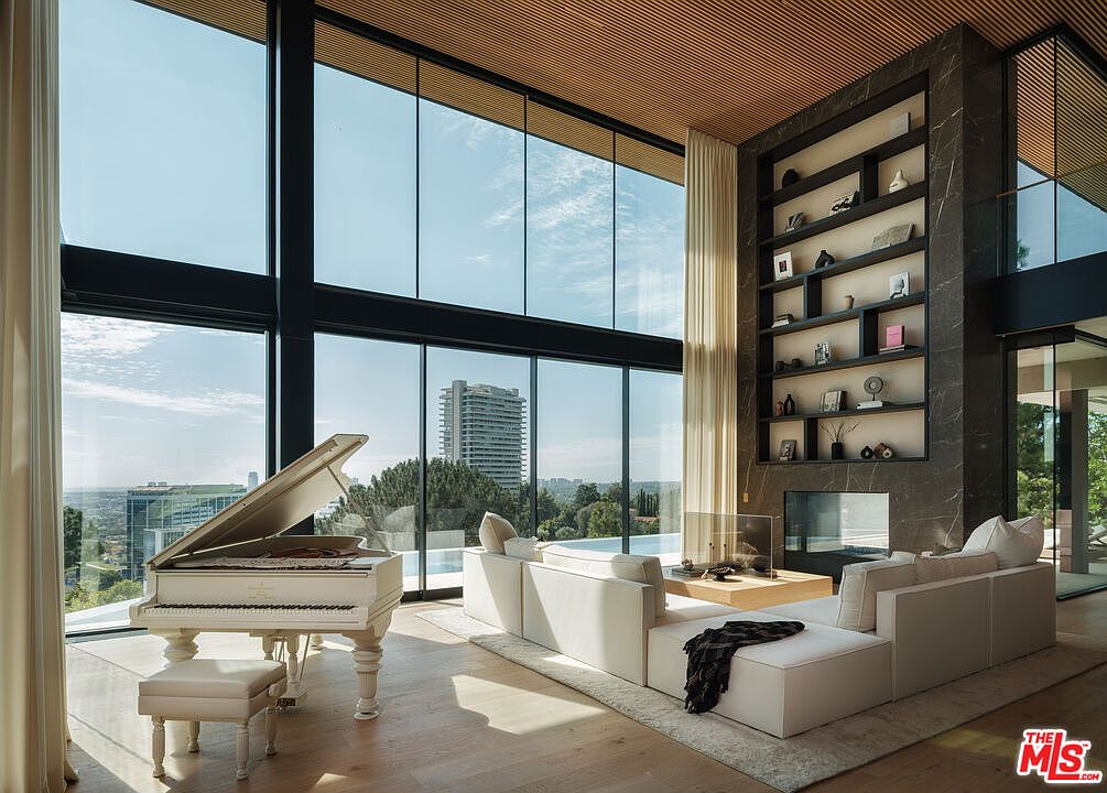 This expansive, double-height living room features floor-to-ceiling glass walls that frame a stunning panoramic city view. The space is anchored by a grand white piano and a large, plush white sectional sofa, complemented by a dramatic dark marble fireplace wall with integrated shelving. The combination of warm wood-slat ceilings and modern, minimalist decor creates a sophisticated and luxurious atmosphere.
