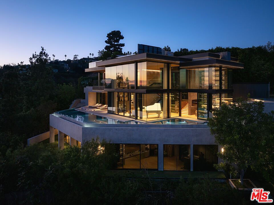 This stunning modern architectural masterpiece is captured at twilight, showcasing a multi-level glass-walled residence perched on a hillside. The home features a sleek, minimalist design with a prominent infinity-edge pool that reflects the evening sky, while warm interior lighting highlights a grand piano and open-concept living spaces. The cinematic perspective emphasizes the seamless integration of luxury indoor-outdoor living against a backdrop of lush, dark foliage and a clear dusk horizon.