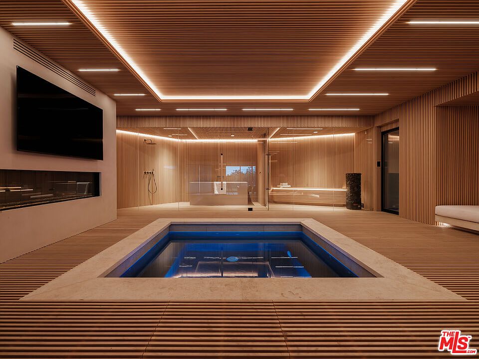 This luxurious indoor spa area features a sunken plunge pool as the central focal point, surrounded by warm, slatted wood paneling that extends from the floor to the ceiling. Behind a glass partition, a sauna and a modern shower setup create a serene, high-end wellness retreat. The space is illuminated by integrated linear LED lighting, creating a sophisticated and tranquil atmosphere perfect for relaxation.